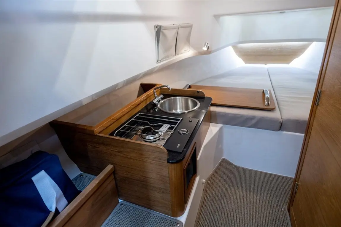 Compact galley in a boat featuring a sink, gas stove, wooden cabinets, and a cushioned sleeping area in the background.